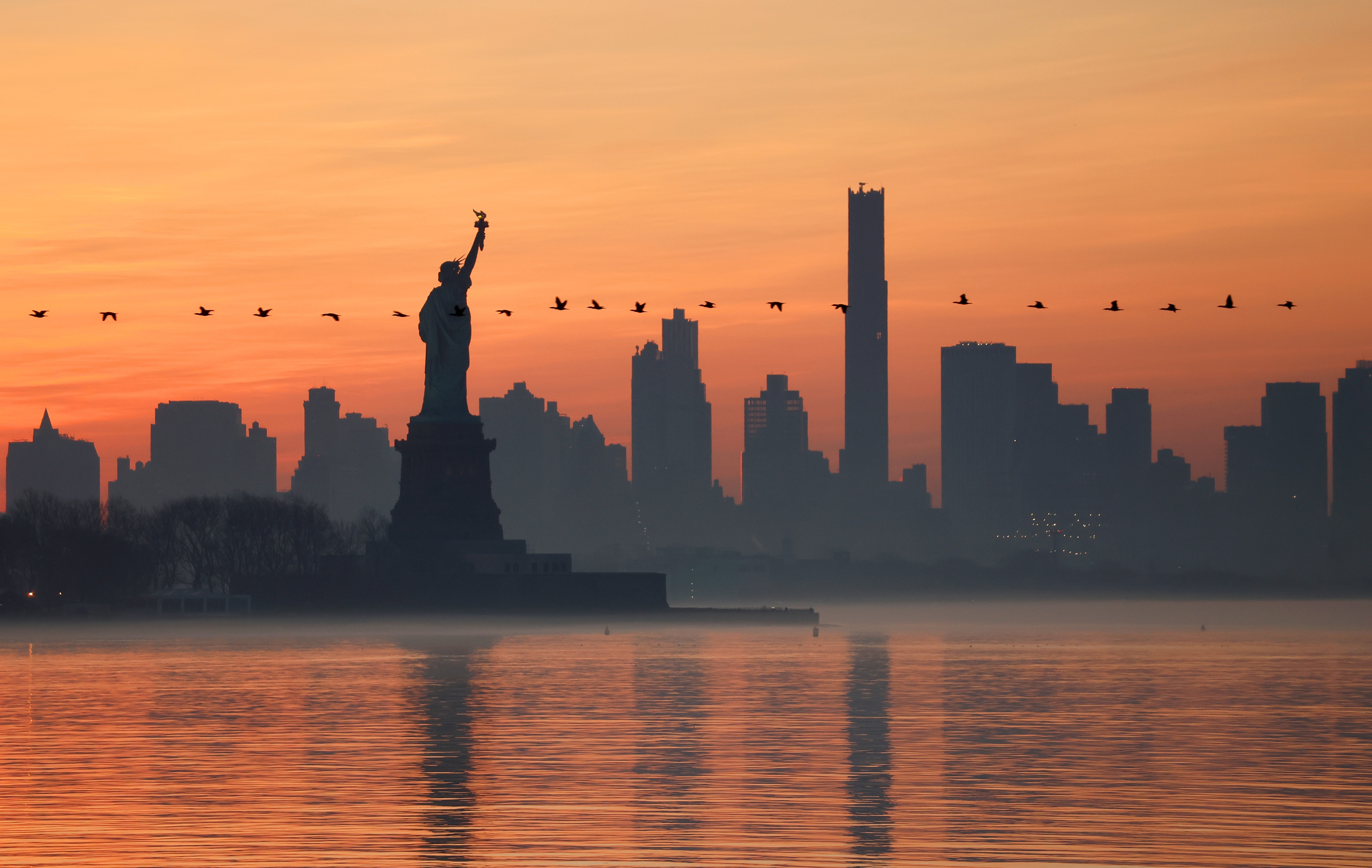 A line of geese flies past the Statue of Liberty and the skyline of Brooklyn at sunrise.