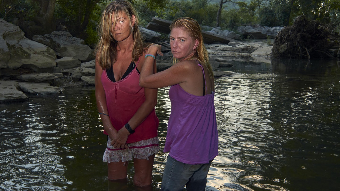 Two women wearing halter tops stand in a river looking straight to camera.