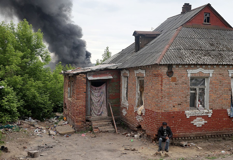 A person sits outside a brick house with a pillar of black smoke rising in the background.