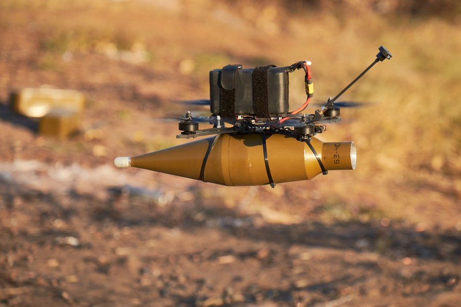 A small four-rotor drone hovers just above the ground, with an explosive strapped beneath it.