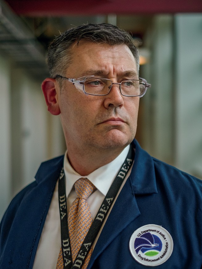 A man wearing glasses, pink tie, white collared shirt, blue jacket with DEA logo, and DEA lanyard