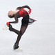A figure skater is photographed mid-turn, leaning back and grasping the blade of the skate on her right foot.