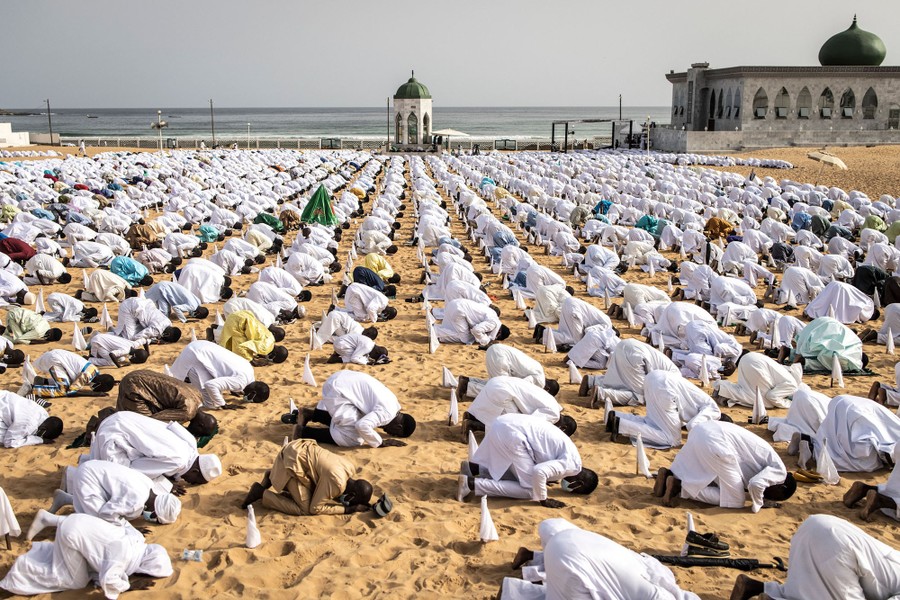 Hundreds of people in white attire kneel together in prayer on a beach.