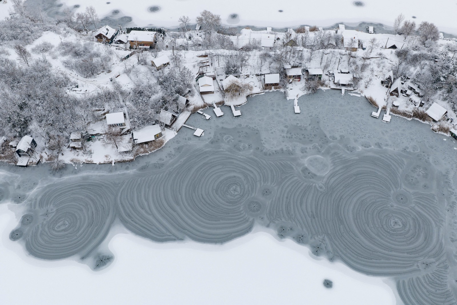 An aerial view of a lakeside neighborhood beside a frozen lake, which has swirling patterns visible in the ice.