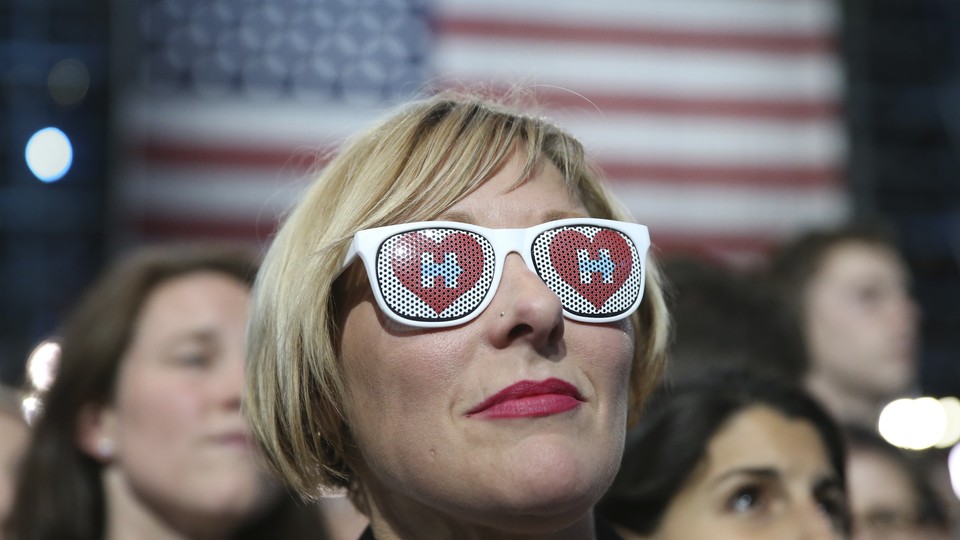 A woman wearing sunglasses with the Clinton campaign logo in front of an American flag