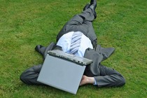 A businessman wearing a suit lays on the grass with a laptop over his face