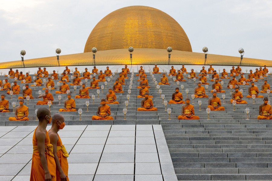 Dozens of monks in orange robes sit cross-legged on the steps of a temple.