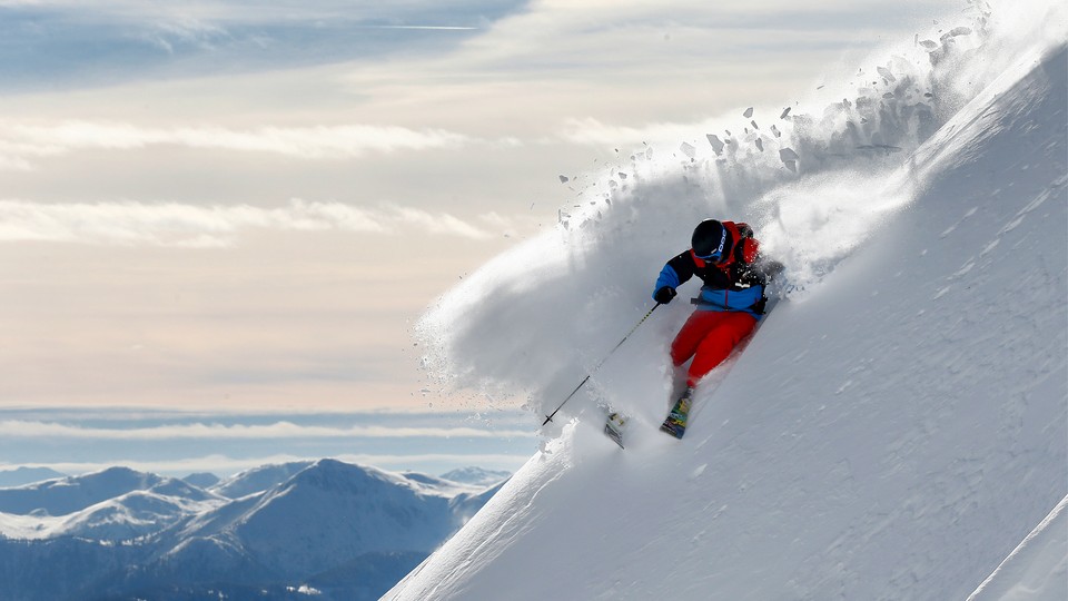 Photograph of a skier in red pants, a blue jacket, and a black helmet carving a turn in deep powder on a steep slope, with mountains in the background