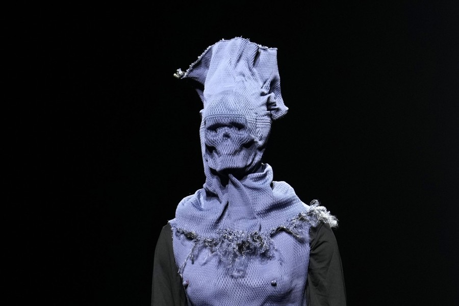 A model presents a garment, topped by a full sheet of cloth wrapped around their head, with a skull-like face.