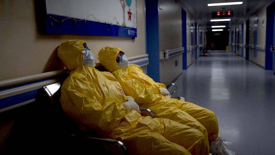 Two hospital workers in yellow hazmat suits rest on chairs in a dimly lit hallway