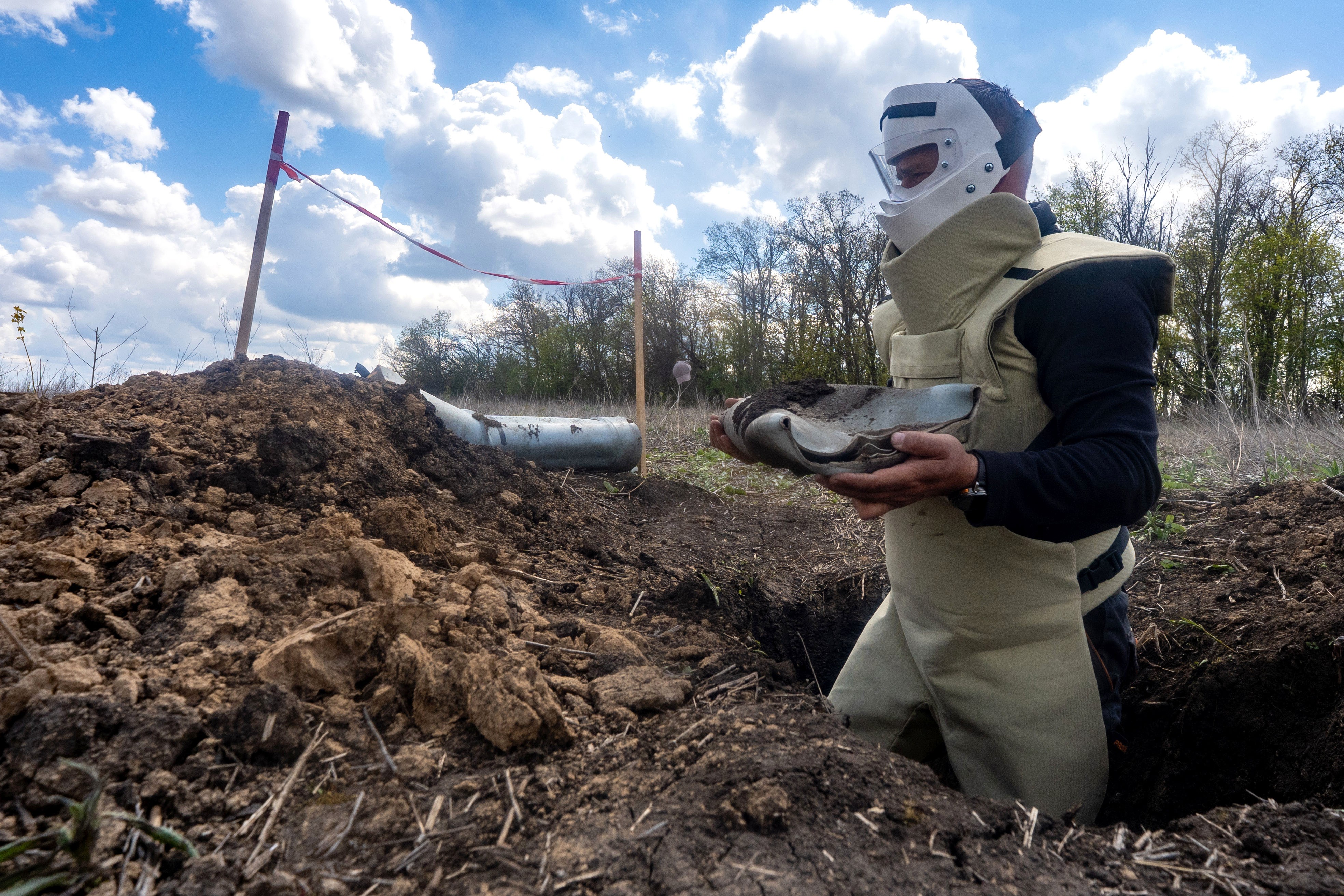 A person wearing protective gear and a face mask removes potential explosive material from a hole in a farm field.