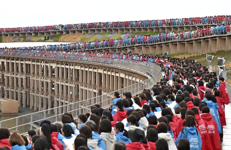A large crowd wearing red, white, or blue raincoats lines up on a tall ring structure, singing.