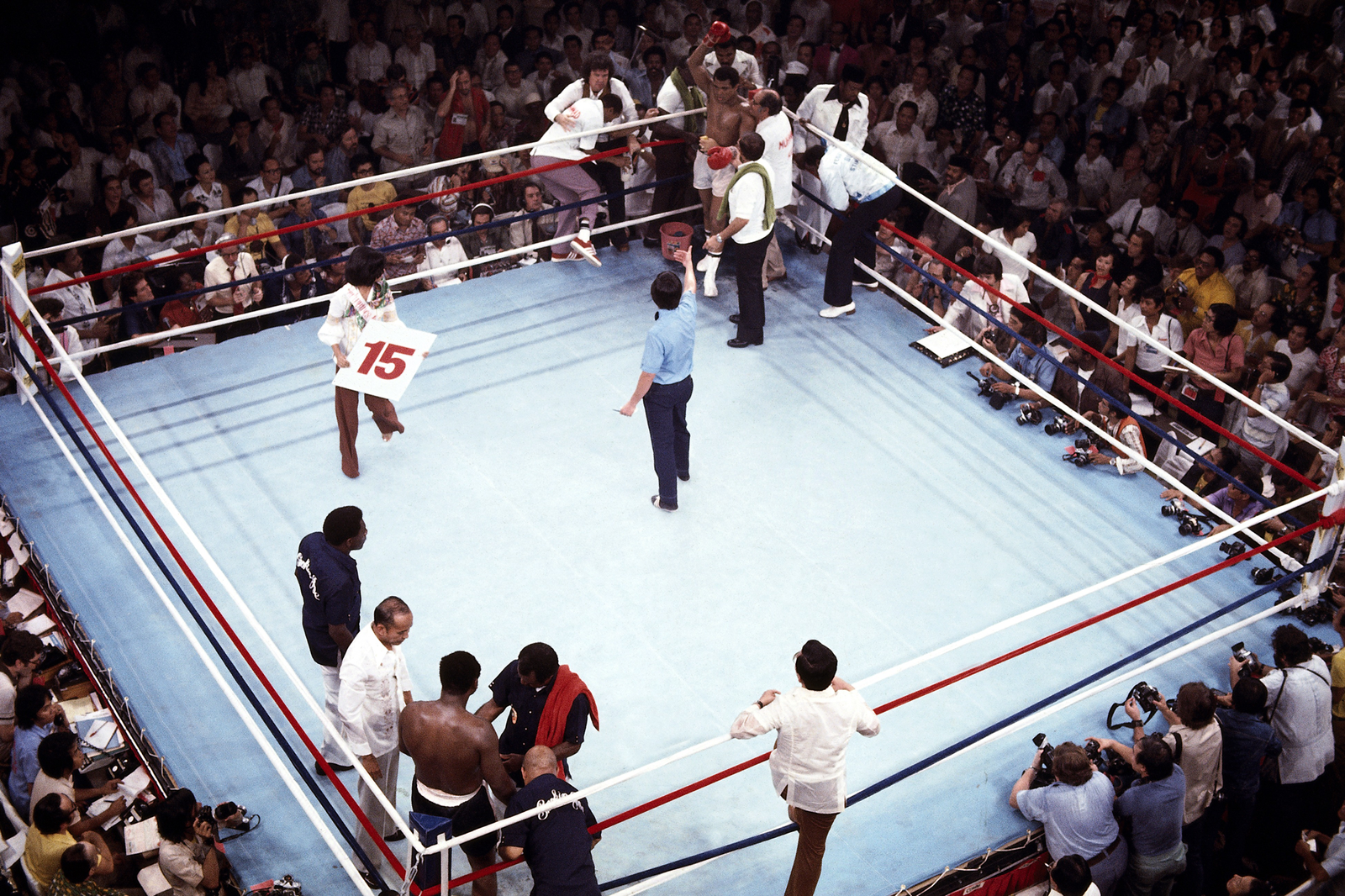 color photo from above of ring with fighters in corners and Ali raising one glove, with person carrying a sign with 15 and the referee Padilla gesturing to Ali