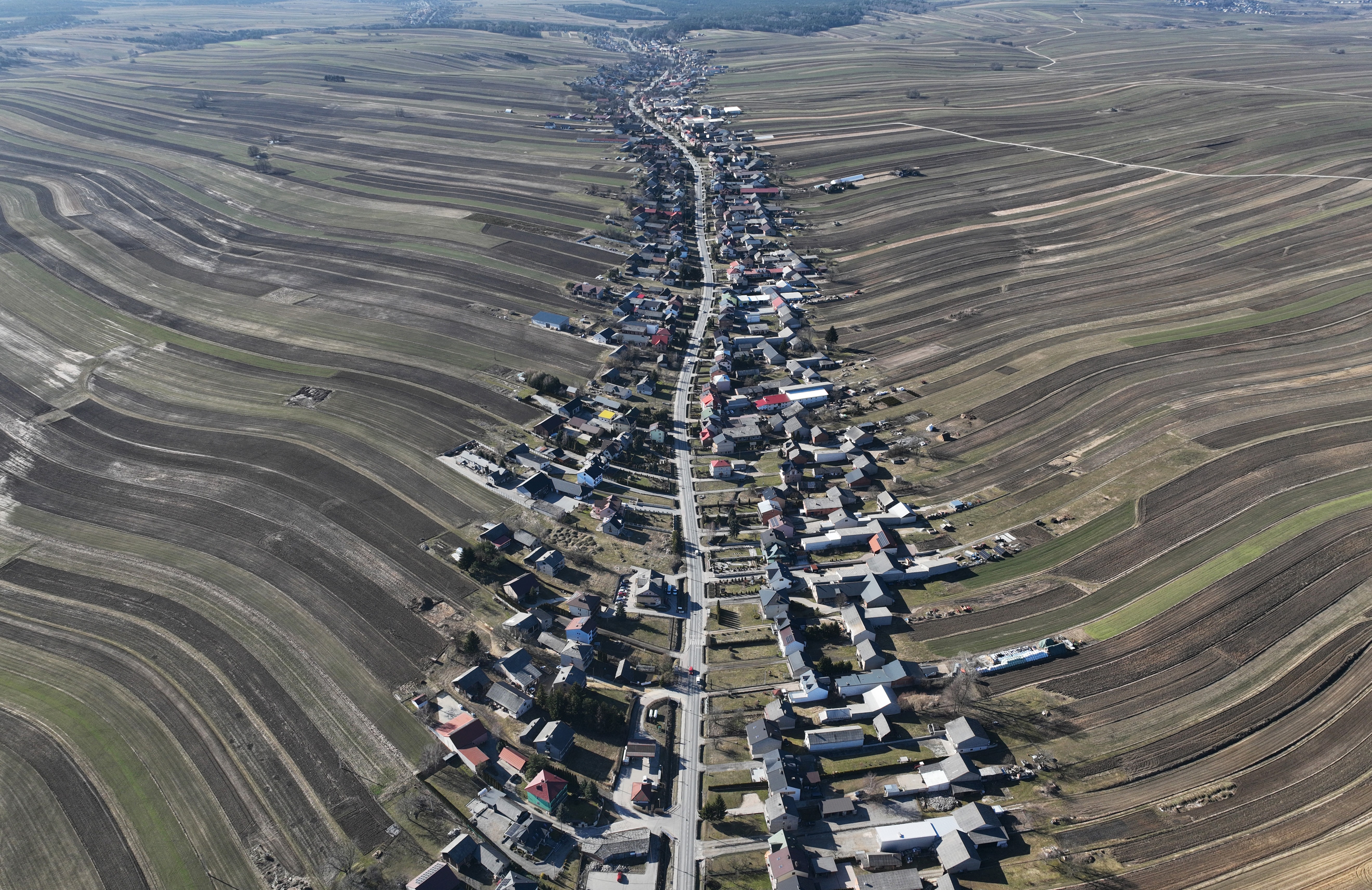 An aerial view of many houses arranged along a single road, surrounded by dozens of long, thin farm plots.