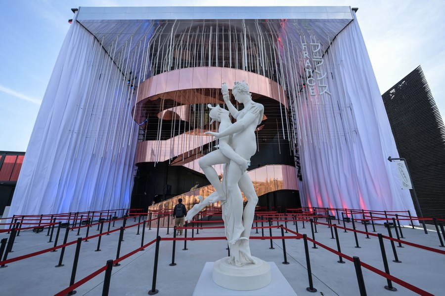 A statue of two intertwined figures stands in front of a large pavilion building with a copper-covered helical staircase in the center.