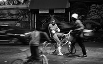 A Black teenager, in focus, sits on a bike with his arms folded, as people and cars around him are blurred.