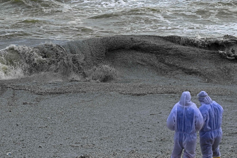 Two Navy members stand on a beach near waves crashing ashore that are thick with plastic debris.