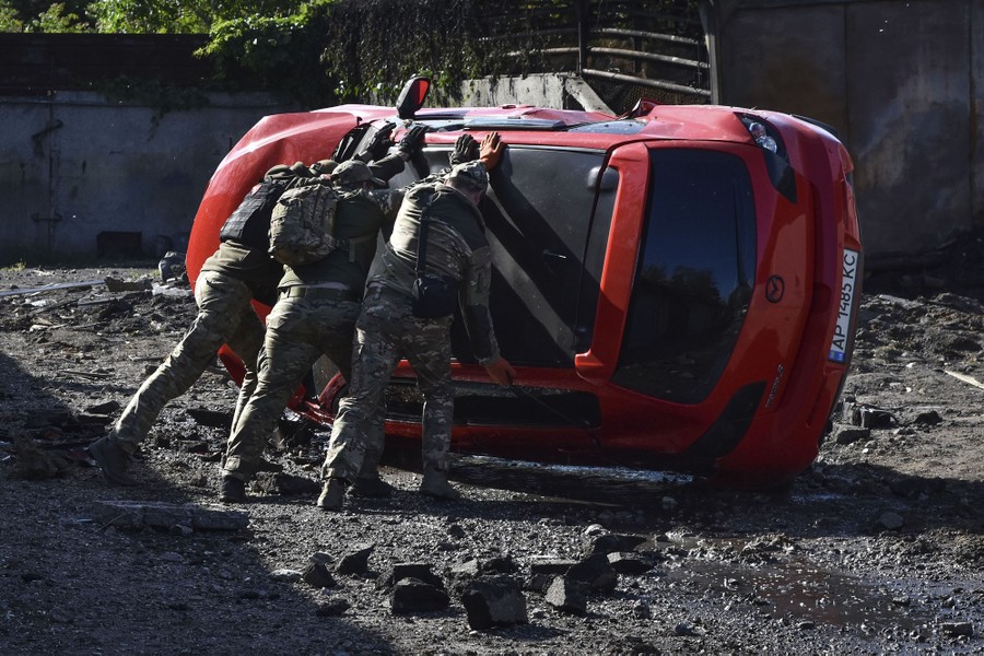 Three soldiers work together to right a car that lies on its side amid scattered rubble.
