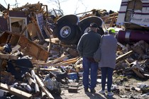 A couple stands close together, surveying extensive damage from a tornado, including a tractor unit of a semitruck atop a pile of debris.