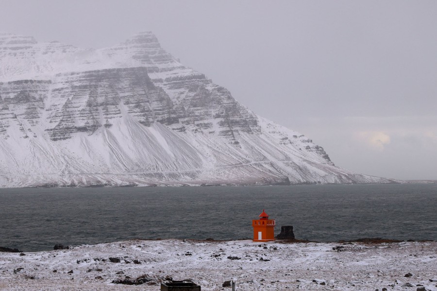 A small red lighthouse stands on a snowy coastline with a mountain visible in the distance.