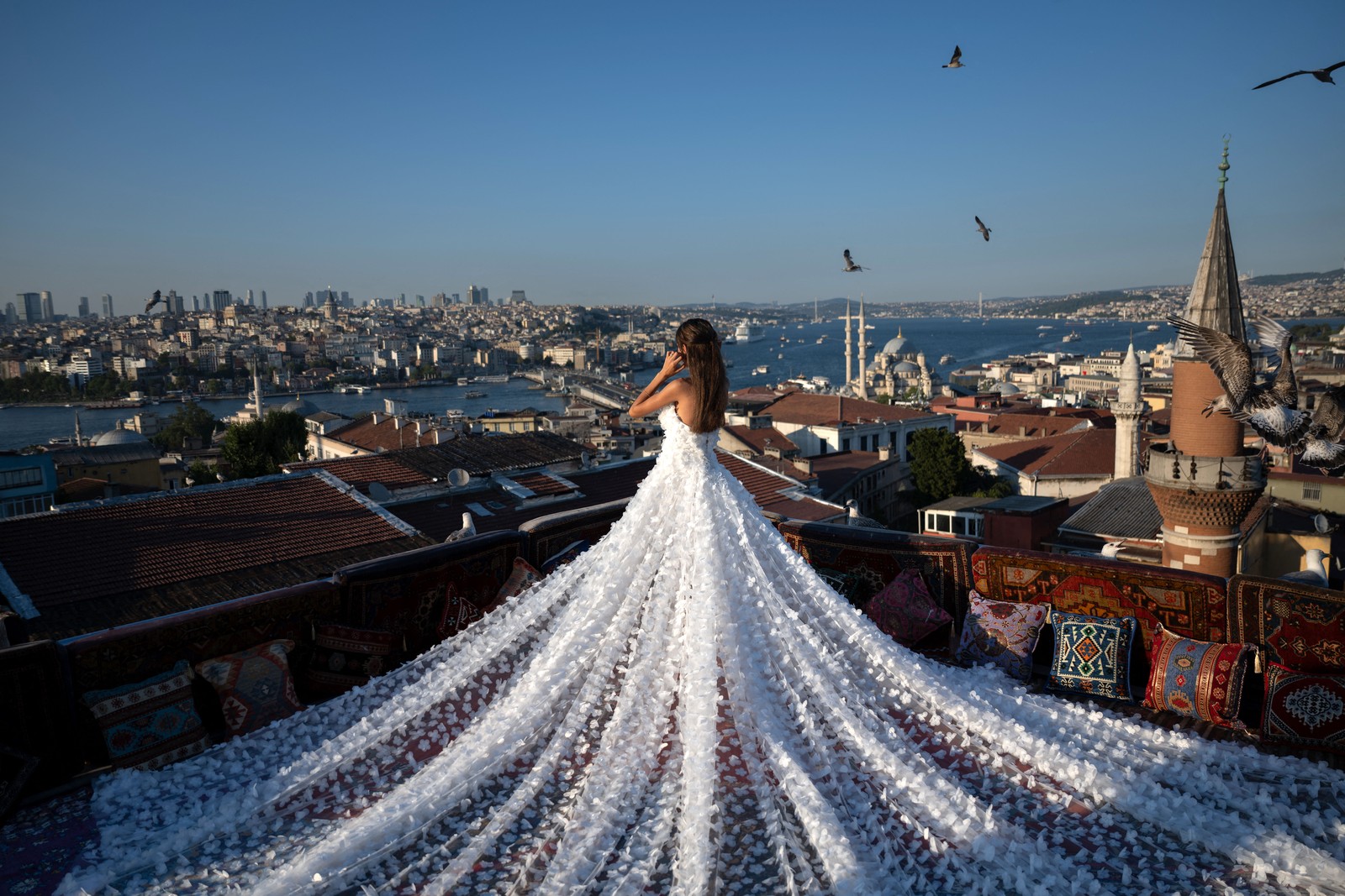 A woman wearing a very long dress stands at the corner of a rooftop overlooking the city of Istanbul.