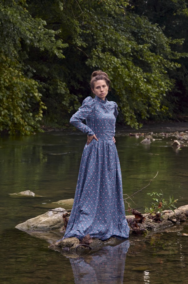The photographer standing on rocks in a river wearing Victorian blue gown. 