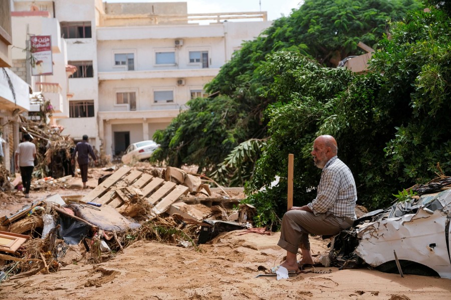 A man sits on a damaged car among mud and debris, in a street.