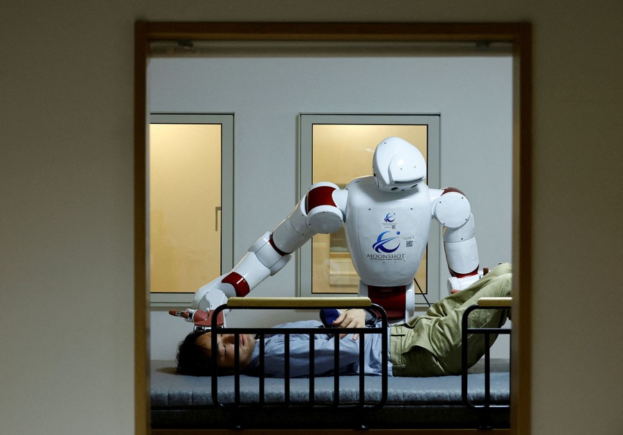 A humanoid robot prepares to roll a human patient onto their side on a bed.