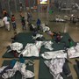 Kids lie on mats, some under foil sheets, on a floor in a detention-center pen.