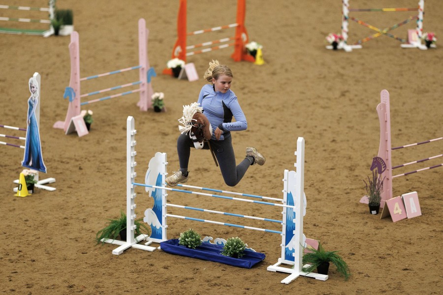 A young person leaps over an obstacle while holding a hobbyhorse.