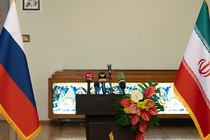 A color photograph of Iranian and Russian flags flanking a podium mounted with several microphones.