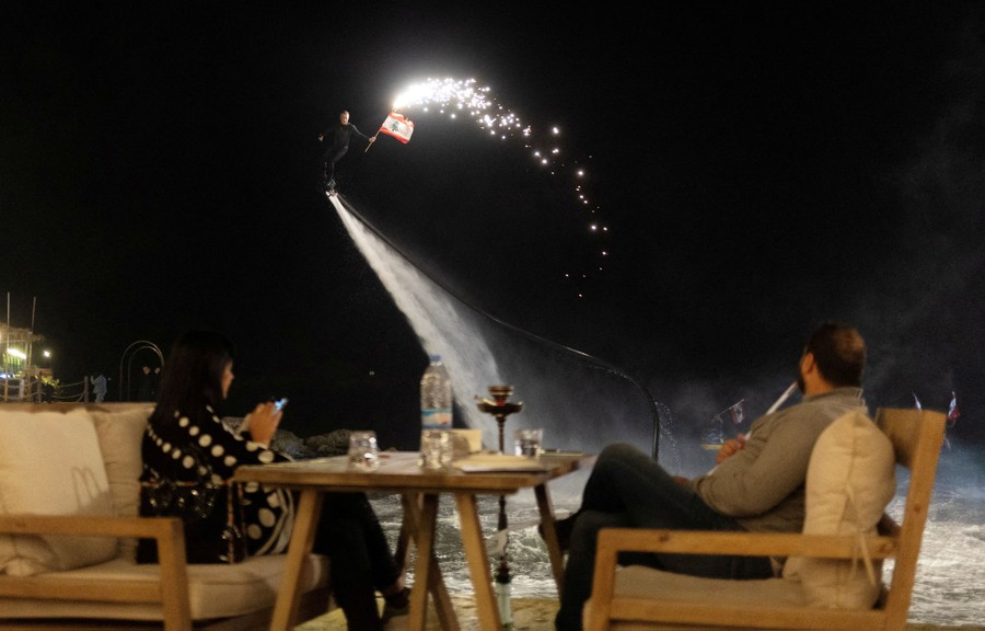 A person rides a water-jet-powered flyboard near diners at a restaurant while carrying a Lebanese flag and fireworks.
