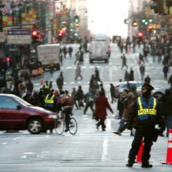 A police officer blocks traffic as pedestrians, bikers, cars, and trucks negotiate Fifth Avenue in New York.