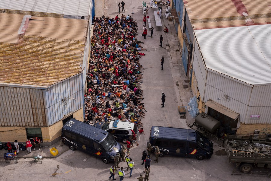 More than 100 minors sit close together in a street beside a building, under the watch of police officers.