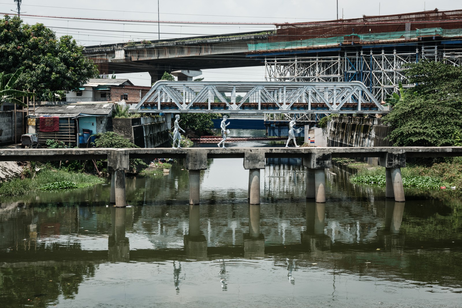 Three silver-painted street performers walk on a pedestrian bridge across a river in an industrial area.