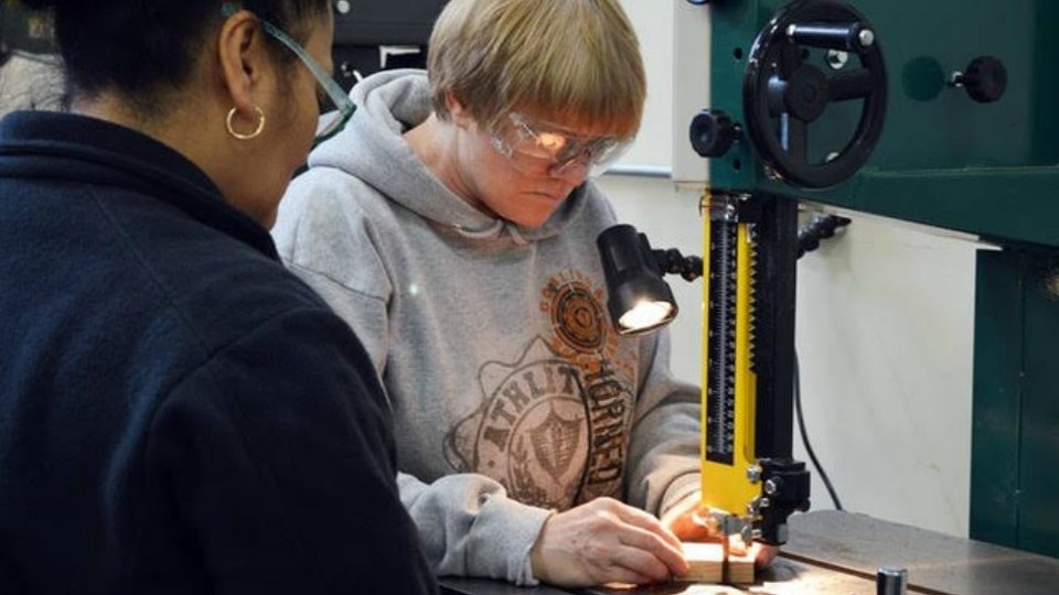 A woman in a sweatshirt and goggles moves a piece of wood through a machine.