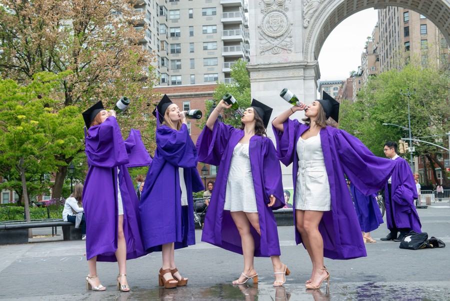 Four young women drink champagne in a park while wearing graduation caps and gowns.