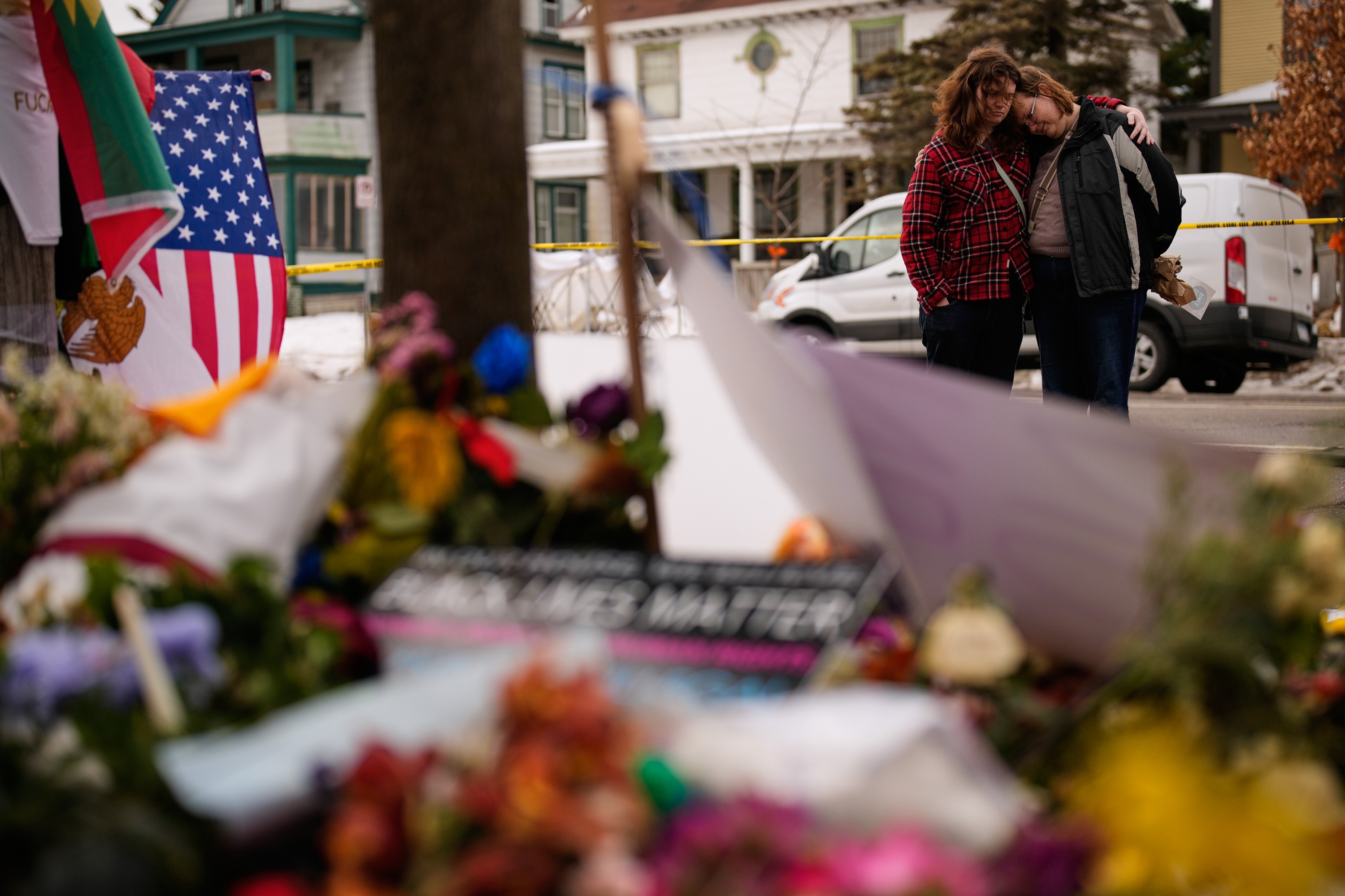 Two people embrace while standing on a street looking at a makeshift memorial filled with flower bouquets.