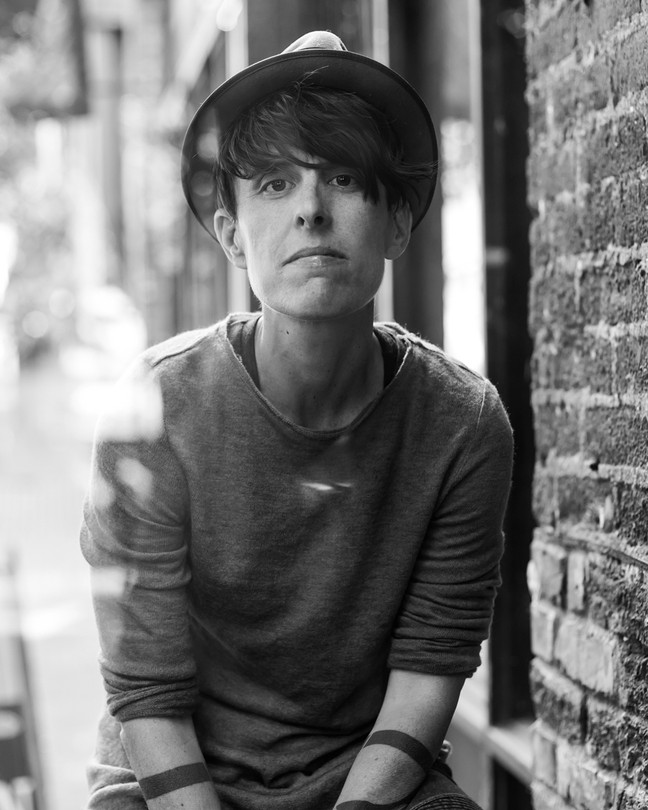 A black-and-white photo of a person with dark hair wearing a narrow hat and a thin sweater beside a brick wall