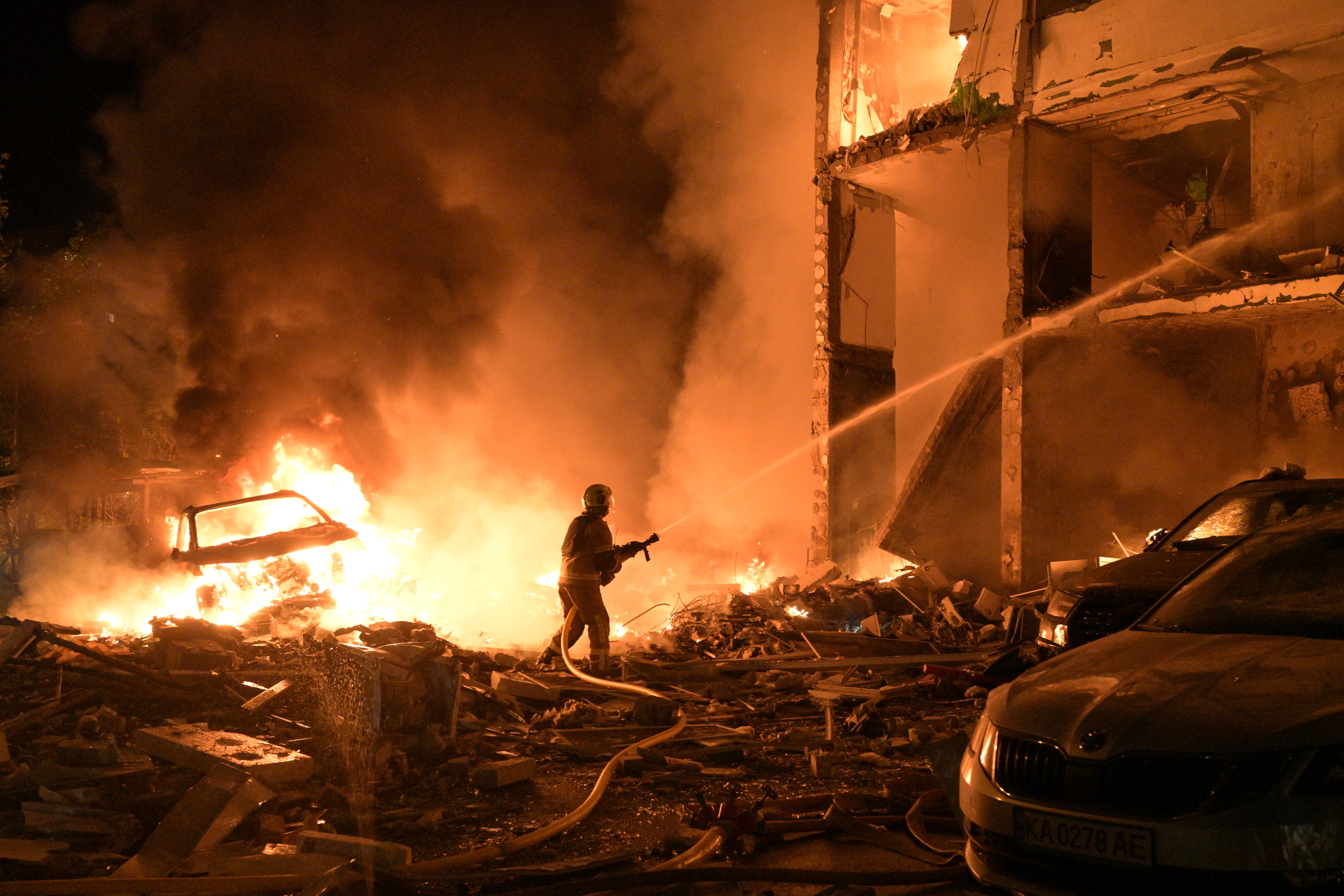 A firefighter stands amid debris from a damaged building, spraying water toward numerous fires in and around the building.