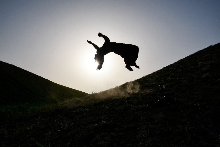 A person performs a backflip on a hillside, seen in mid-air, silhouetted against the sun.