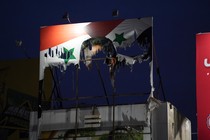 A tattered and torn billboard of the Syrian flag with Bashar al-Assad’s face on it lit up atop a building at night
