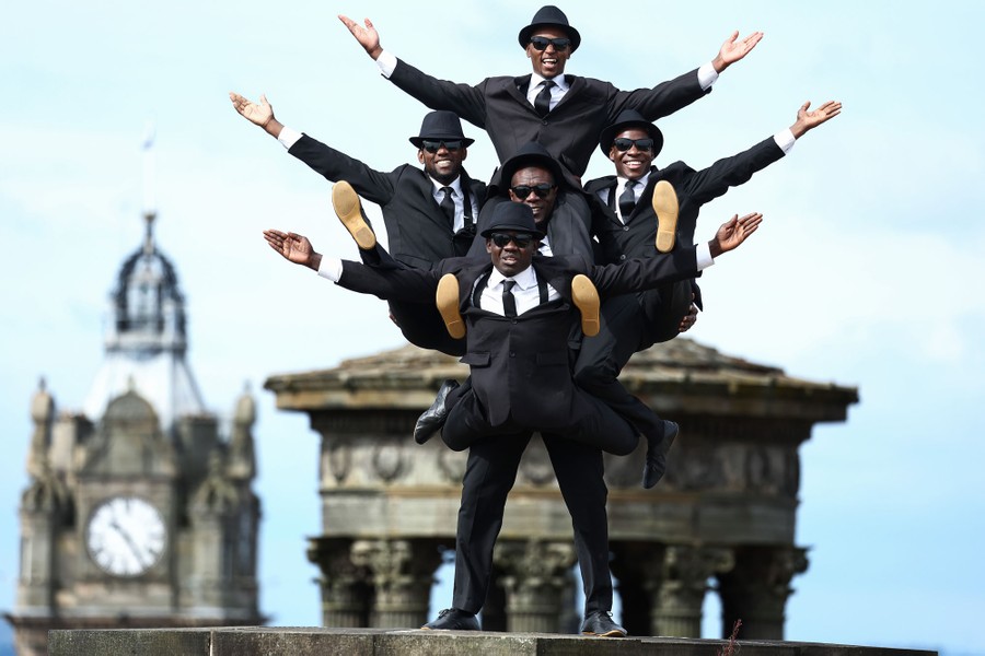 A group of performers in black suits pose—four of them perched gymnastically on the back of one man.