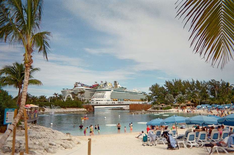 photo of sheltered sandy beach with palms, umbrellas, and chairs with two large docked cruise ships in background