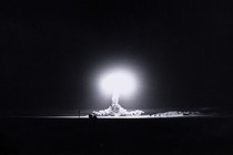Black-and-white photo of a pitch-black night sky with brilliant white glowing nuclear cloud in distance at center.