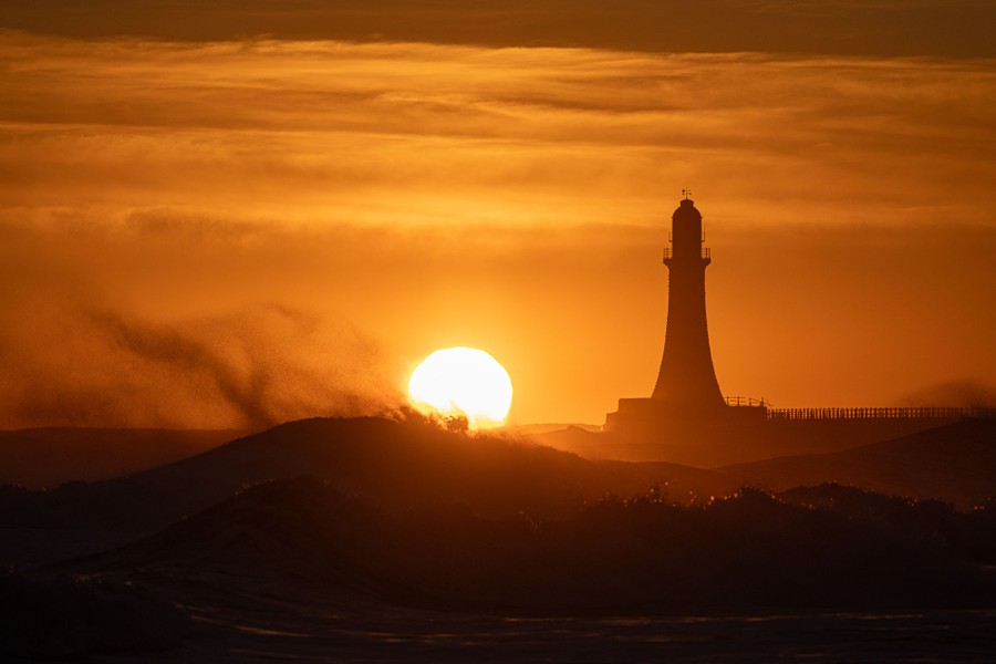 The sun rises past waves and a lighthouse.
