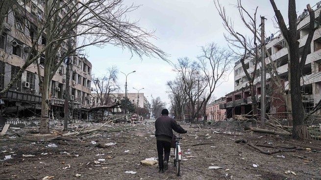 A man wearing all black walks with a bike in the middle of a road which is surrounded with debris from the war.