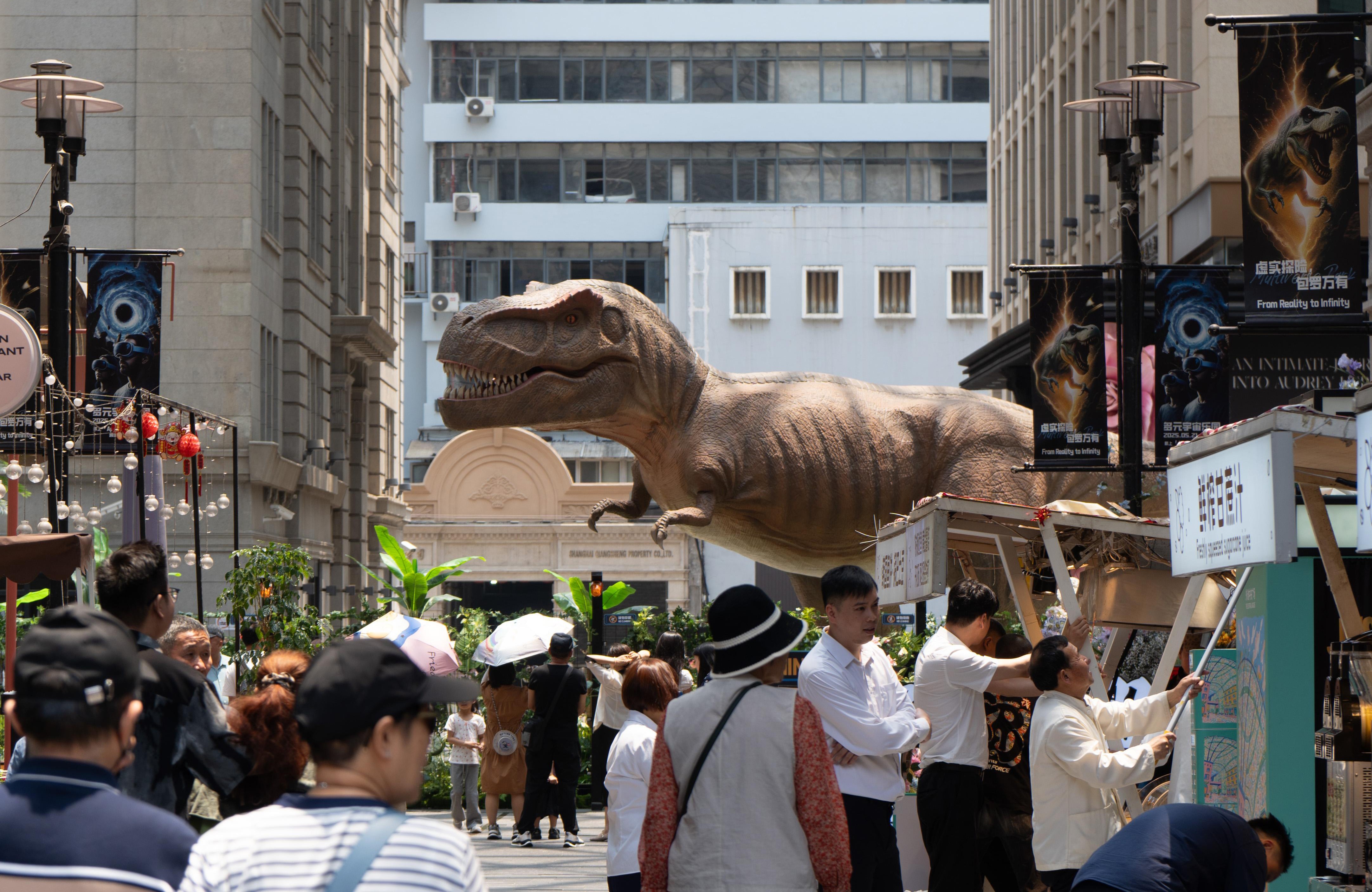 People walked through an open market with a large dinosaur statue behind it.