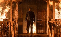 A masked Michael Myers stands on the porch of a burning house