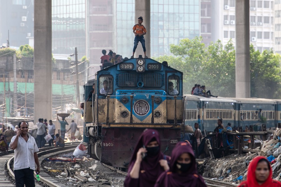 A boy stands on top of the locomotive of a passenger train, with other riders visible on the train's roof behind him, as the train moves through an area crowded with people.